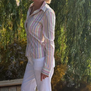 Randig pastellskjorta i bomull - Säljer en långärmad skjorta i tunna pastellfärger med ränder i rosa, blått, grönt, gult och lila. Skjortan har klassisk krage, knäppning framtill och två bröstfickor. Materialet är lätt och känns svalt, perfekt för varmare dagar.