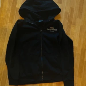 Peak Performance hoodie - Svart hoodie från Peak Performance med dragkedja och huva. Klassisk logga broderad på bröstet. Mjuk insida och två fickor framtill. Perfekt för chill dagar eller när du vill ha en enkel och snygg look. Ett litet hål där fram