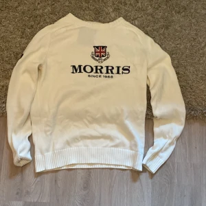 Vit stickad tröja från Morris - Snygg vit stickad tröja från Morris med broderad logga och brittisk flagga på bröstet. Tröjan har långa ärmar, ribbade muddar och en svart siffra på ena ärmen. Perfekt för dig som gillar klassisk och stilren design.