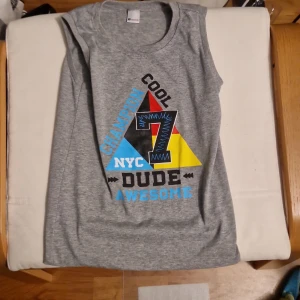 Grå ärmlös t-shirt Assign, tryck - Grå ärmlös t-shirt från Assign med färgglatt tryck framtill. Trycket har text som 'CHAMPION', 'COOL', 'NYC', 'DUDE', 'AWESOME' och siffran 7 i blått, rött, gult och svart. Perfekt för en avslappnad och sportig look.