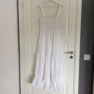 Vit långklänning från Zara - Superfin vit långklänning från Zara med smock upptill och breda axelband. Klänningen har volang nertill och är tillverkad i ett lätt och luftigt material, perfekt för varma dagar. Enkel och stilren design som ger en fräsch look.