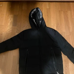 Svart Moncler dunjacka med huva - Svart dunjacka från Moncler med huva och tydlig logga på ärmen. Jackan har dragkedja framtill, två fickor med dragkedja och Moncler-tryck på insidan av huvan. Perfekt för kalla dagar och riktigt snygg streetstil.