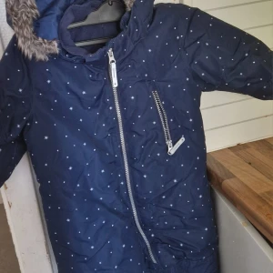 Mörkblå vinterjacka med stjärnmönster - Mysig mörkblå vinterjacka för 2-5 åringar med vitt stjärnmönster och fuskpäls på huvan. Jackan har snedställd dragkedja framtill och en extra dragkedja på bröstet. Perfekt för kalla dagar och riktigt snygg design!