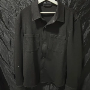 Svart overshirt från Zara - Stilren svart overshirt från Zara med klassisk krage och två stora bröstfickor. Skjortan har tryckknappar framtill och är tillverkad i ett mjukt och stretchigt material som känns lite tjockare. Perfekt att slänga över en t-shirt för en cool look. Passar bra till nästan allting och perfekt inför vår och sommar. Men funkar även fint till vintern med en hoodie under. Storlek M skick 9,5/10.