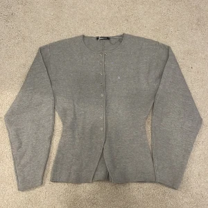 Grå kofta från Gina Tricot - Säljer en grå kofta från Gina Tricot med lång ärm och rund halsringning. Koftan har tryckknappar hela vägen framtill och en snygg croppad passform med lätt utsvängd nederkant. Perfekt att styla med kjol eller jeans för en clean look.💗