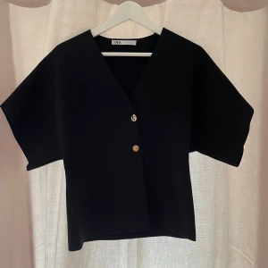 Svart blus med guldknappar från zara - Svart blus från Zara med v-ringning och två snygga guldknappar framtill. Modellen är väldigt stil ren!