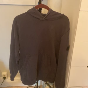 Mörkgrå hoodie från Stone Island Junior - Säljer en mörkgrå hoodie från Stone Island Junior med klassisk logotyp-patch på vänster ärm. Tröjan har huva, magficka och långa ärmar. Perfekt för dig som gillar streetwear och vill ha en clean look med snygga detaljer.