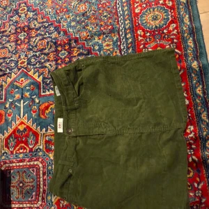 Olivgrön manchesterkjol från Flashlights - Säljer en olivgrön kortkjol i manchester från Flashlights. Kjolen har klassiska fickor fram och bak, knapp och dragkedja framtill samt cool retrovibe. Perfekt för dig som gillar utilitystil och vill ha något som sticker ut i garderoben.