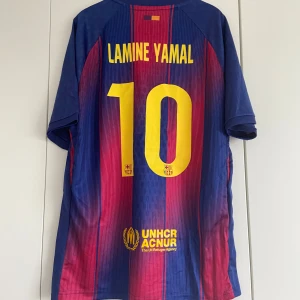 FC Barcelona Lamine Yamal tröja - Snygg FC Barcelona matchtröja med Lamine Yamal och nummer 10 på ryggen. Tröjan har klassiska blå och röda vertikala ränder, gula tryck och UNHCR-logga. Kortärmad modell i lätt och ventilerande material, perfekt för fotbollsälskare.