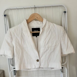 Vit croppad kavaj från Zara - Snygg vit croppad kavaj från Zara med korta ärmar och klassisk krage. Kavajen har en knapp framtill och en liten bröstficka. Tillverkad i linne för en lätt och fräsch känsla, perfekt för varmare dagar.