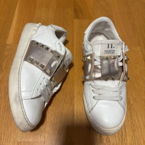 Valentino Rockstud sneakers vit/guld - Säljer ett par vita Valentino Rockstud sneakers i skinn med bred metallicband i champagnefärg och ikoniska nitar. Skorna har snörning, platt sula och rund tå. Perfekta för dig som vill sticka ut med en lyxig och edgy vibe.