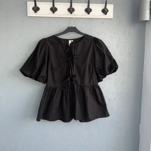Svart blus med puffärm från Nelly - Svart blus från Nelly med kort puffärm och snygga knytband framtill. Modellen har en lätt utställd passform och rund halsringning. Tillverkad i bomull för en skön känsla och luftig look. Perfekt för dig som gillar trendiga detaljer och volym i ärmarna. Knappast använd så toppen är i mycket bra skick🤍🌸