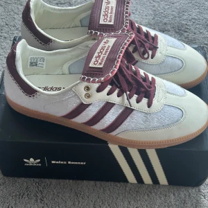 Adidas Wales Bonner Samba sneakers - Unika Adidas Wales Bonner Samba sneakers i vitt och vinrött med klassiska tre ränder och kontrasterande sömmar. Ovandel i skinn och textil med vinröda snören och detaljer. Snygg gummisula och retrovibbar, perfekt för dig som vill sticka ut.