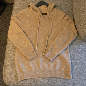 Beige hoodie i kashmir med snörning - Mjuk beige hoodie i kashmir med långa ärmar och huva. Tröjan har snörning med metalländar och ribbade muddar vid ärmslut och nederkant. Perfekt för dig som gillar chill och stilren look.