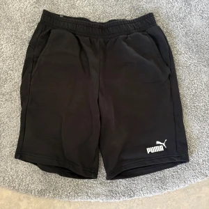 Svarta shorts från Puma - Svarta shorts från Puma med elastisk midja och två sidofickor. Klassisk och enkel design med Puma-logga i vitt på ena benet. Perfekta för träning eller chill. Mjukt material som känns skönt mot huden.