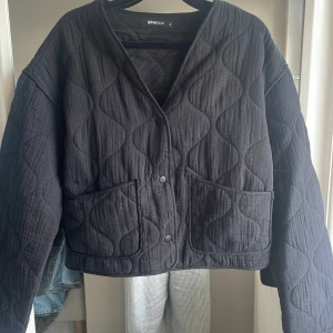 Svart quiltad jacka Gina Tricot S - Quiltad svart jacka från Gina Tricot i storlek S. Jackan har oversized passform, stora fickor framtill och tryckknappar. Snygg vågig quiltning och V-ringad hals. Perfekt för lager-på-lager och en cool vardagslook.