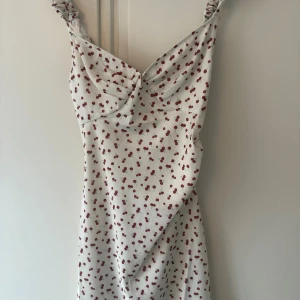 Vit kortklänning med körsbär från Adoore - Supersöt vit kortklänning från Adoore med röda körsbärsmönster. Klänningen har smala axelband med twistad detalj och en snygg v-ringning framtill. Perfekt för sommarens alla tillfällen och ger en fräsch och lekfull vibe. Denna går inte längre att få tag på så den är one of a kind. 