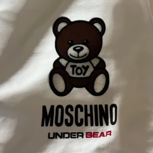Vit Moschino tröja med björntryck - Säljer en vit tröja från Moschino med det ikoniska björntrycket på bröstet. Tröjan har rund halsringning och är tillverkad i mjuk bomull. Perfekt statement-plagg för dig som gillar lekfull design och streetstyle. Riktigt skön o snygg tröja, använd 2 gånger.