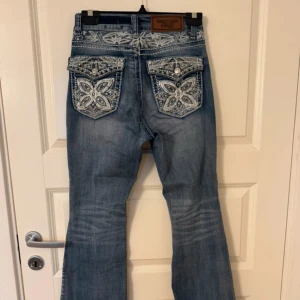 Blå bootcut jeans Westfield Eagle - Snygga blå bootcut jeans från Westfield Eagle med broderade detaljer och paljetter på bakfickorna. Jeansen har låg midja, dekorativa sömmar och glittriga knappar. Perfekt för dig som gillar jeans med extra bling och unik stil.