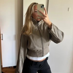 Beige skinnjacka med hög krage - Snygg beige skinnjacka med hög ribbad krage och dragkedja framtill. Jackan har en croppad och loose passform med långa ärmar och ribbade detaljer vid midjan och ärmslut. Använd 1 gång så den är i nyskick!! Köpt för 999kr