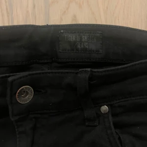 Svarta jeans från Tiger of Sweden - Svarta jeans från Tiger of Sweden med klassisk femficksdesign och smal passform. Jeansen har en stilren look med svarta sömmar och metallknapp framtill. Perfekta för dig som gillar en clean och enkel stil.
