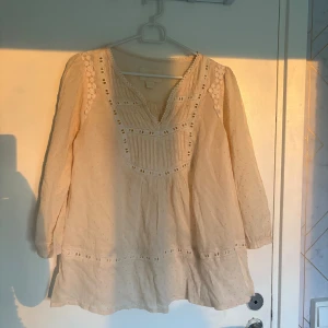 Beige blus med spets från H&M - Säljer en beige blus från H&M Collection med v-ringning och dekorativa spetsdetaljer längs axlar och bröst. Blusen har lång ärm, små prickar i tyget och broderade detaljer. Perfekt för dig som gillar romantisk och boho stil.