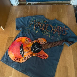 Alice In Chains handmålad t-shirt - Unik handmålad t-shirt med Alice In Chains-tema. Stor gitarr i orange och rött på ryggen, coola texter och detaljerade illustrationer på framsidan. Basfärgen är mörkgrå/blå. Perfekt för dig som vill sticka ut och älskar grunge och konstnärliga plagg.