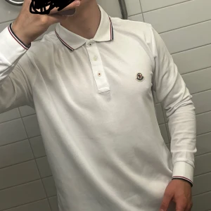 Vit långärmad pikétröja från Moncler - Vit långärmad pikétröja från Moncler med klassisk krage och knäppning framtill. Tröjan har Moncler-logga på bröstet och snygga rödblå ränder på krage och ärmslut. Tillverkad i högkvalitativ bomull för en skön och stilren känsla. Tröjan är använd fåtal gånger och i mycket gott skick, alltså som ny. Modellen på bilden är ca 179 cm lång och väger ca 75kg. Tröjan är storlek L och säljes då den tyvärr är lite i största laget och där med inte används. Nypris: 3900kr Pris kan diskuteras vid snabb affär
