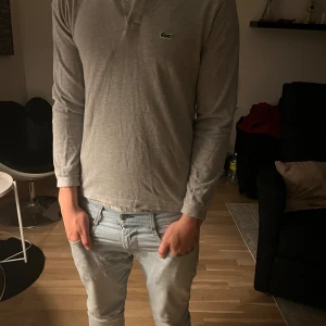 Lacoste - Säljer en stilren grå långärmad pikétröja från Lacoste med klassisk krage och knäppning framtill. Tröjan har det ikoniska gröna krokodilmärket broderat på bröstet och är tillverkad i mjuk bomull. Perfekt för dig som gillar clean och enkel stil.