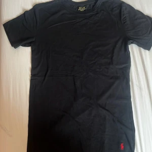 Svart t-shirt från Polo Ralph Lauren - Svart t-shirt från Polo Ralph Lauren med klassisk röd broderad logga vid nederkanten. Modellen är slim fit och har korta ärmar. Tillverkad i mjuk bomull som känns skön mot huden. Perfekt för dig som gillar stilrena och tidlösa plagg.