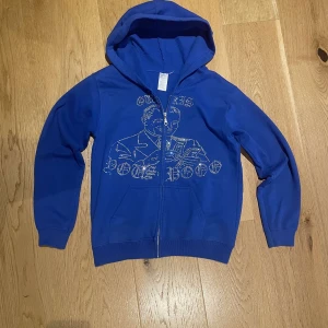 Blå hoodie med strasstryck och huva - Blå hoodie med dragkedja och huva. Framsidan har ett stort motiv av en person och text i glittrande strasstenar. Tröjan har långa ärmar, två fickor fram och ribbade muddar. Perfekt för dig som gillar statement-plagg och coola detaljer.