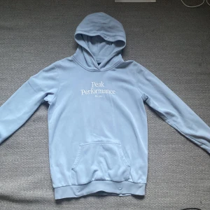Ljusblå hoodie från Peak Performance - Snygg ljusblå hoodie från Peak Performance med vit logga på bröstet. Klassisk känguruficka framtill och huva. Perfekt för en chill och avslappnad stil. Mjuk insida och ribbade muddar vid ärmslut och nederkant. Pris kan diskuteras! 