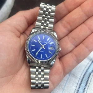 Blå Shaarms klocka med länkarmband - Stilren klocka från Shaarms med blå urtavla och datumvisning. Klockan har ett klassiskt silverfärgat länkarmband i metall och tydliga indexmarkeringar. Perfekt accessoar för dig som gillar en clean och modern look.