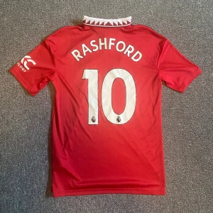 Manchester United kit 2022/23 - Säljer en officiell Manchester United fotbollströja från Adidas med Rashford och nummer 10 på ryggen. Tröjan är röd med svarta detaljer, vit krage med rött mönster och klubbmärke på bröstet. Materialet är lätt och andas, perfekt för match eller träning. S/M