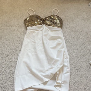 Vit klänning med guldpaljetter - Supersnygg vit kortklänning med smala axelband och glittrig överdel i guldpaljetter. Kjolen har omlott-design och slits framtill för extra vibe. Perfekt för dig som vill sticka ut med en glam look. Materialet känns mjukt och följsamt mot huden.