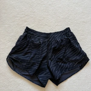 Svarta zebramönstrade shorts Lululemon - Snygga svarta träningsshorts från Lululemon med zebramönster. De har elastisk midja och är tillverkade i återvunnen polyester och elastan, vilket gör dem både lätta och bekväma. Perfekta för dig som vill sticka ut på gymmet eller löprundan.