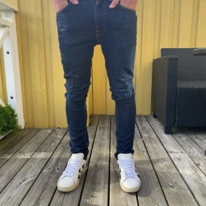 Nudie Jeans - Säljer ett par mörkblå skinny jeans från Nudie Jeans i storlek W29 L32. Jeansen har klassiska orangea sömmar, fem fickor och en tight passform som sitter snyggt längs benen. Tillverkade i stretchig denim för extra komfort.