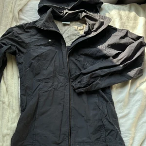 Mörkblå vindjacka från Columbia S - Säljer en mörkblå vindjacka från Columbia med huva och dragkedja framtill. Jackan har två knappar vid bröstet, elastiska muddar och dragsko nertill. Lättviktig och perfekt för blåsiga dagar. Snygg och enkel design som funkar till det mesta.