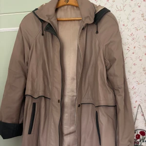 Beige parkajacka med huva, stl L - Säljer en beige parkajacka i storlek L med svarta detaljer och dragkedja. Jackan har huva, långa ärmar och två fickor framtill. Materialet är mjukt och känns som syntet, perfekt för kyligare dagar. Snygg och enkel design med svarta kontrastdetaljer. Hjälper min mormor, använd ett fåtal gånger💗vet ej märke