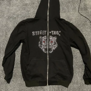 Svart hoodie med tigrar och strass - Svart hoodie med huva och dragkedja från Street Tag. Framsidan har ett stort tryck med två tigrar i strass och röda ögon samt texten 'STREET TAG'. Mjukt material och två fickor framtill. Perfekt för en cool streetstyle-look.
