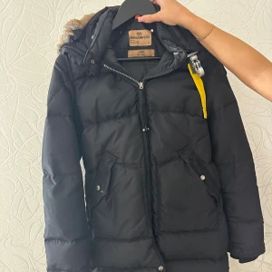 Svart dunjacka Parajumpers med päls - Svart dunjacka från Parajumpers med fluffig päls på huvan och gul detalj vid halsen. Jackan har dragkedja, två fickor med knappar och en varm, quiltad design. Perfekt för kalla dagar med snygg och lyxig look.