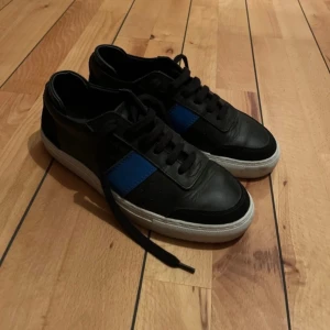 Axel Arigato svarta sneakers med blå detalj - Snygga svarta sneakers från Axel Arigato med en blå panel på sidan och vit sula. Skorna är i skinn och har klassisk snörning. Perfekta för dig som gillar stilrena och moderna sneakers med en färgklick.