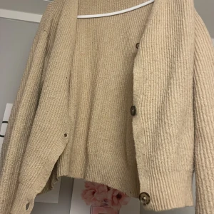 Beige stickad kofta från 157 - Mysig beige stickad kofta från 157 med v-ringning och stora knappar framtill. Koftan har ribbad struktur och långa ärmar, perfekt för lager på lager. Enkel och stilren design som passar till det mesta.