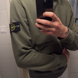 Stone Island grön hoodie med patch - Säljer en grön hoodie från Stone Island med klassisk svart och gul patch på vänster ärm. Tröjan har huva med snörning och en stor magficka. Mjuk insida och ribbade muddar vid ärmslut och nederkant. Perfekt för dig som gillar streetwear.