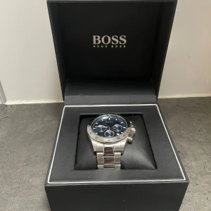 Hugo Boss silverfärgad klocka - Stilren klocka från Hugo Boss med blå urtavla och silverfärgat armband i metall. Klockan är en kronograf och går som den ska. Bra skick och sparsamt använd. 