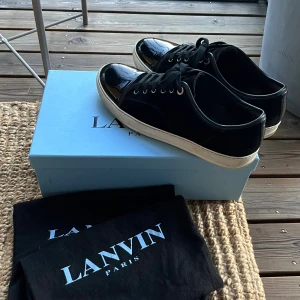 Lanvin Cap Toe - Snygga svarta sneakers från Lanvin i mocka och med lackad tå. Kommer med originalkartong och dustbags. Perfekta för dig som gillar stilrena och lyxiga sneakers. En liten defekt (bild 5) på sulan, höger sko, smutset på yttersulan kommer inte av.  Ingen original innersula. Skorna tagits hand om extremt väl och är i väldigt bra skick. Storlek 44 EU.
