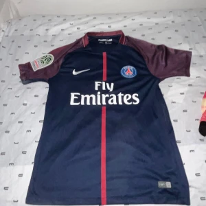PSG matchtröja Nike S - Paris Saint-Germain fotbollströja från Nike i marinblått med vinröda ärmar och röd rand mitt på. Broderat PSG-märke på bröstet, Dri-FIT-material och Fly Emirates-tryck. Klassisk rund hals och korta ärmar. Perfekt för dig som älskar fotboll och PSG.