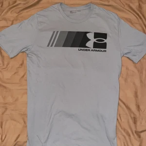 Under Armour t-shirt - Snygg ljusgrå t-shirt från Under Armour med svart och grått grafiskt tryck på bröstet. Modellen är loose fit och har korta ärmar. Perfekt för dig som gillar sportig och avslappnad stil. Tröjan är nästan helt ny.