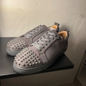 Gråa Louboutin sneakers med nitar - Säljer ett par gråa sneakers från Christian Louboutin i mocka med coola silverfärgade nitar på tån. Skorna har klassisk röd sula, rund tå och snörning. Insidan är beige och sulan är i gummi. Perfekta för dig som vill sticka ut med en edgy vibe.
