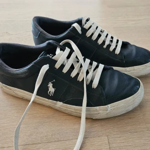 Mörkblåa sneakers från Polo Ralph Lauren - Snygga mörkblåa sneakers från Polo Ralph Lauren med vit logga på sidan och vita snören. Skorna har en klassisk låg profil, rund tå och platt sula. Tillverkade i skinn med vit yttersula och Polo tryck under sulan. Perfekta för en stilren och avslappnad look.